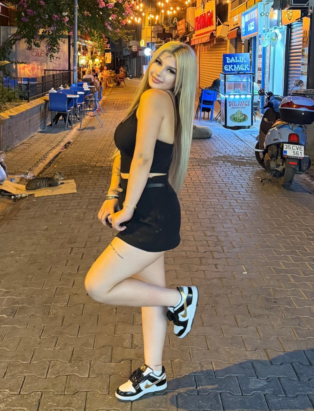 Woman with long blonde hair posing on a lit city street at night, wearing a black tank top, black mini skirt, and white sneakers.