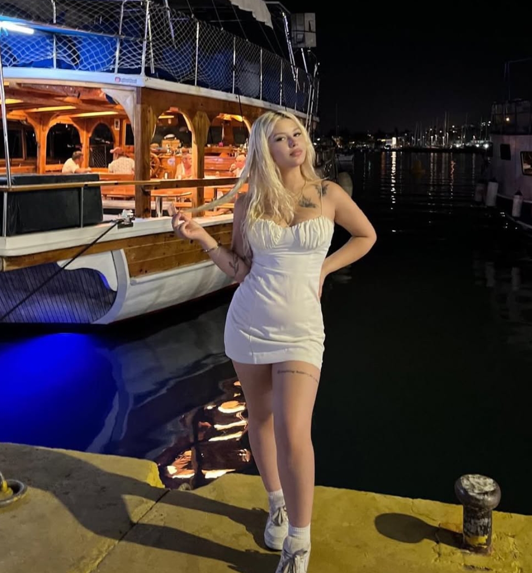 Young woman in a white strapless mini dress and white sneakers poses on a dock at night beside a lit wooden boat.