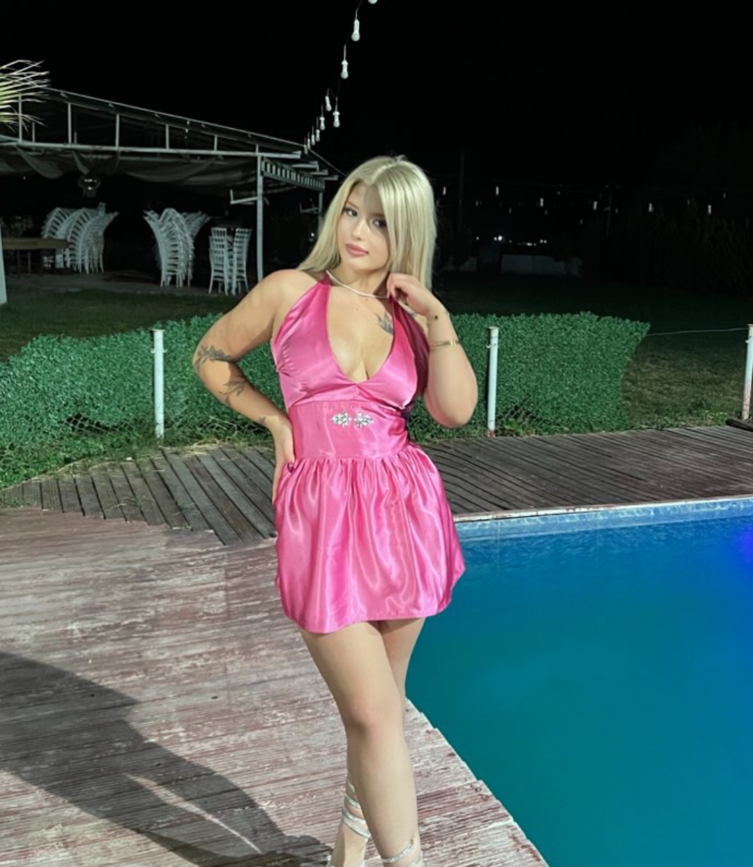 Woman in a pink halter mini dress posing by a pool at night, with string lights overhead.