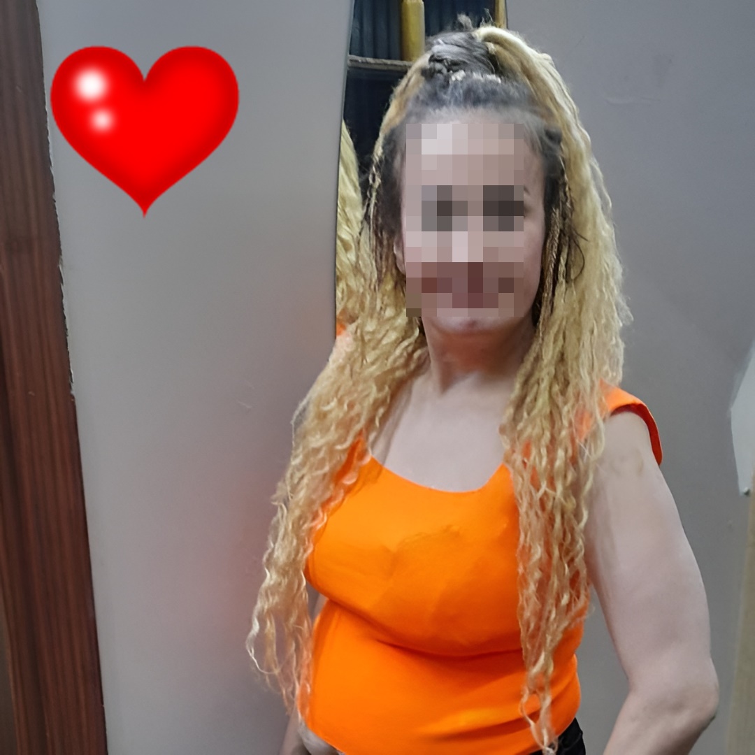 Person with long blonde dreadlocks wearing an orange sleeveless top indoors; face blurred, a red heart emoji to the left.
