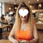 Woman with long curly blonde hair in an orange top sits at a cafe table holding a cup of coffee; face covered by a white heart sticker for privacy.