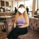 Person with blond braids wearing a lavender tank top sits on a tufted mauve couch in a cafe; their face is covered by a white heart, obscuring identity.