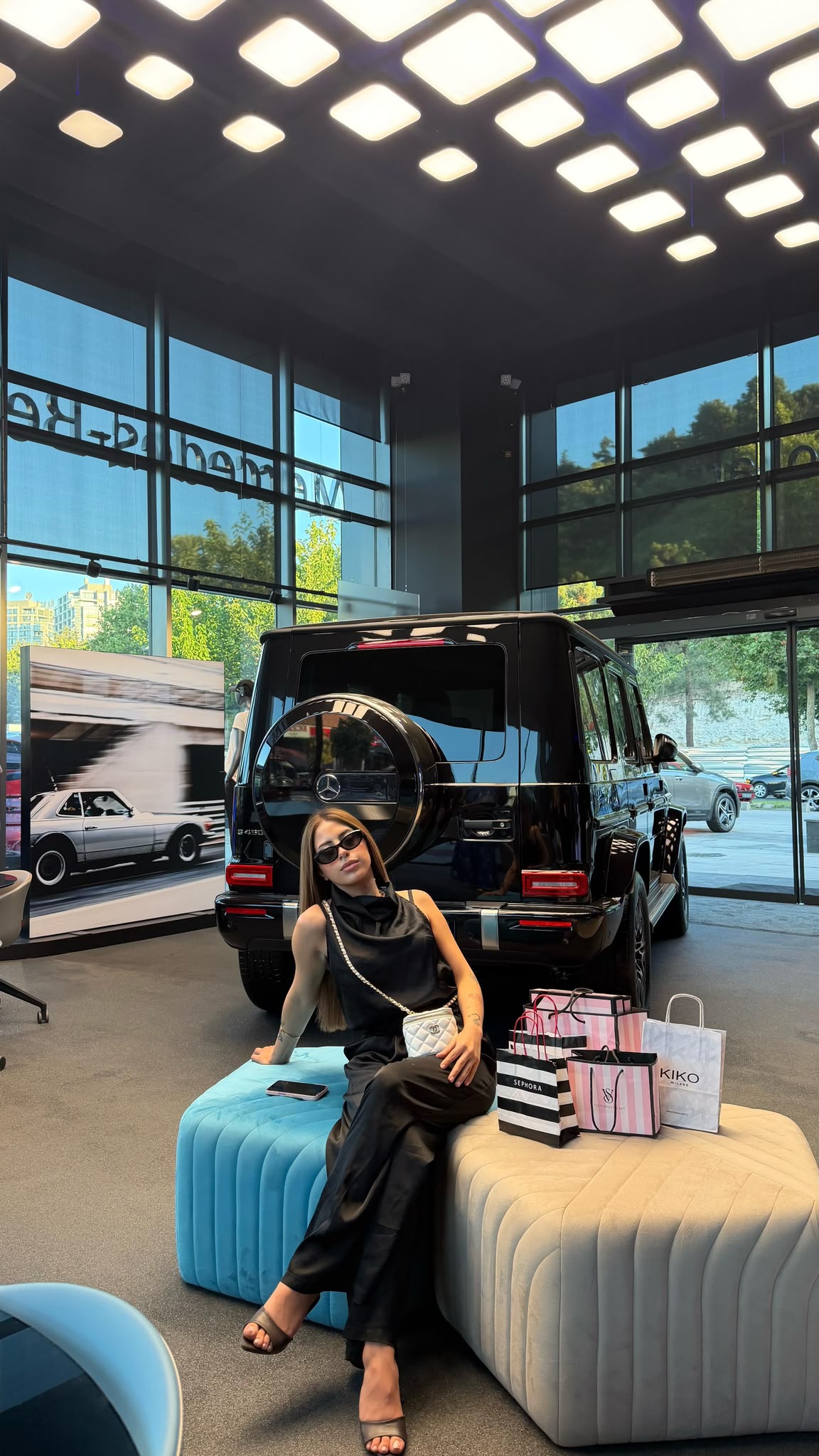 Woman in black outfit and sunglasses sits on blue and beige tufted ottomans in a luxury car showroom, with a black Mercedes SUV and shopping bags nearby.