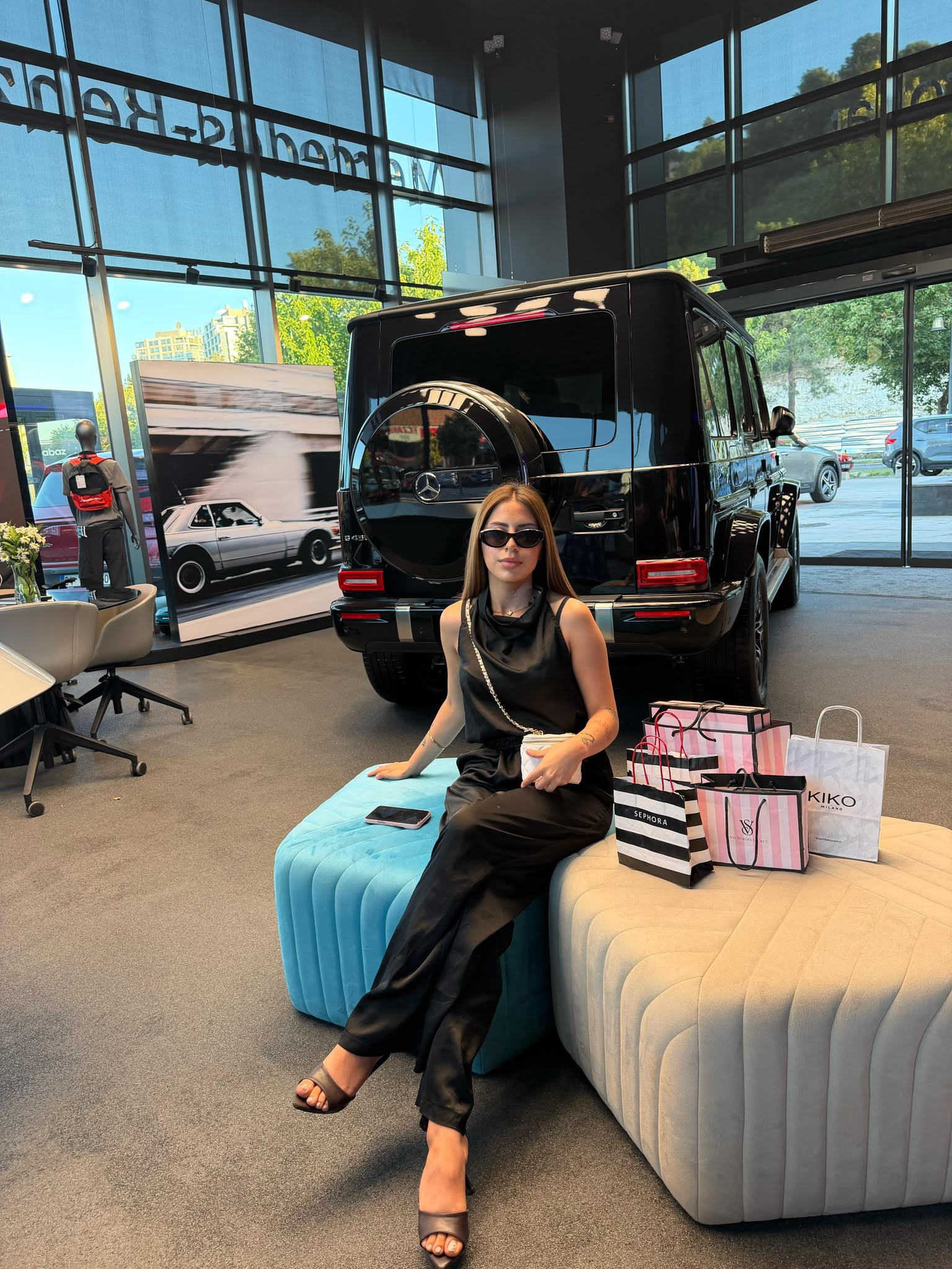 Woman in black dress and sunglasses sits on blue and beige ottomans in a car showroom, with a black luxury SUV and shopping bags nearby.