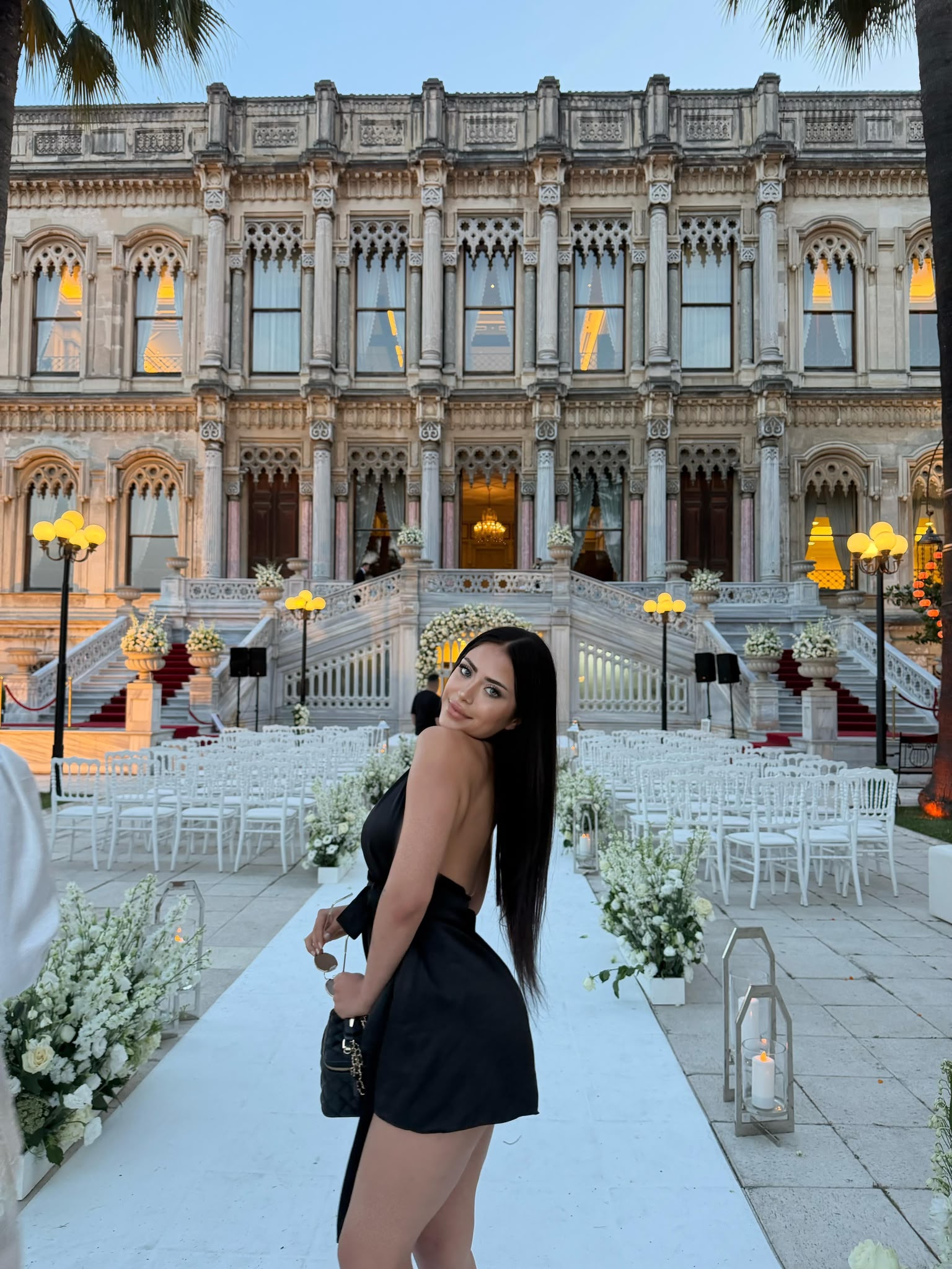 Elegant woman in a black dress posing on a white aisle in front of a grand ornate historical building during a wedding setup with white chairs and floral decor.