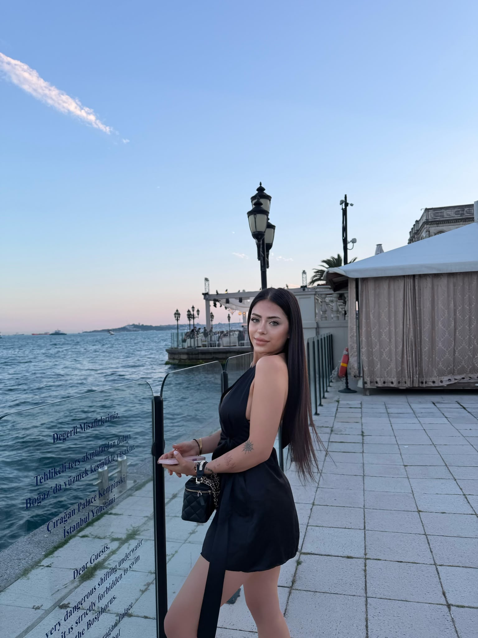 Woman in a black dress posing by a glass railing along a waterfront promenade with the sea and sky behind her.