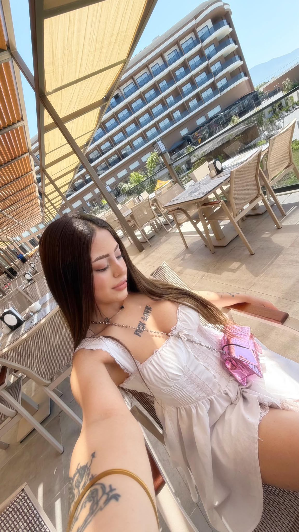 Selfie of a woman in a white dress with tattoos on her arm, taking a sunny outdoor selfie at a cafe patio with modern buildings in the background.