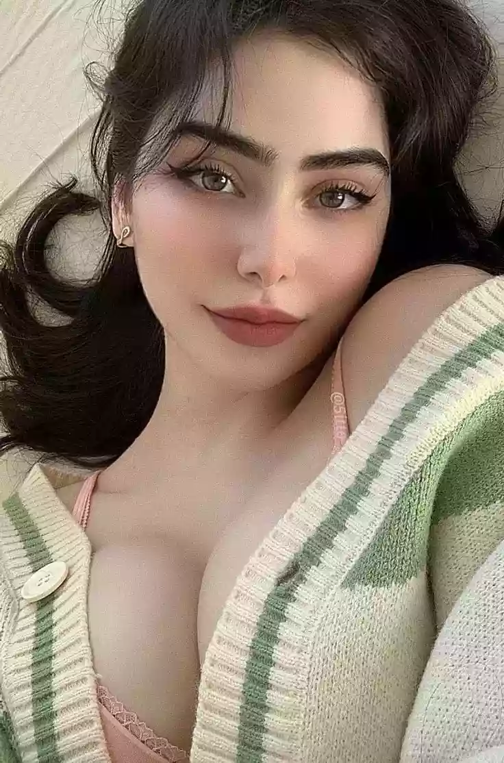 Close-up selfie of a young woman with dark hair and makeup, wearing a pink bra under a striped cardigan, looking at the camera.